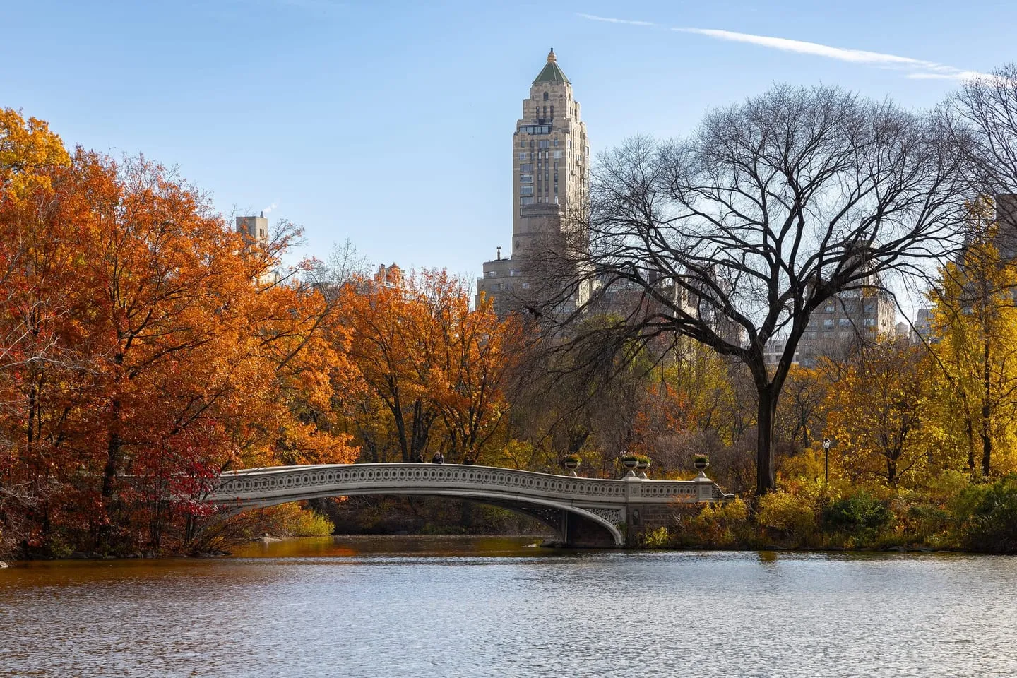 Bow Bridge crossing the Lake in Central Park among autumn trees, with the San Remo towers behind