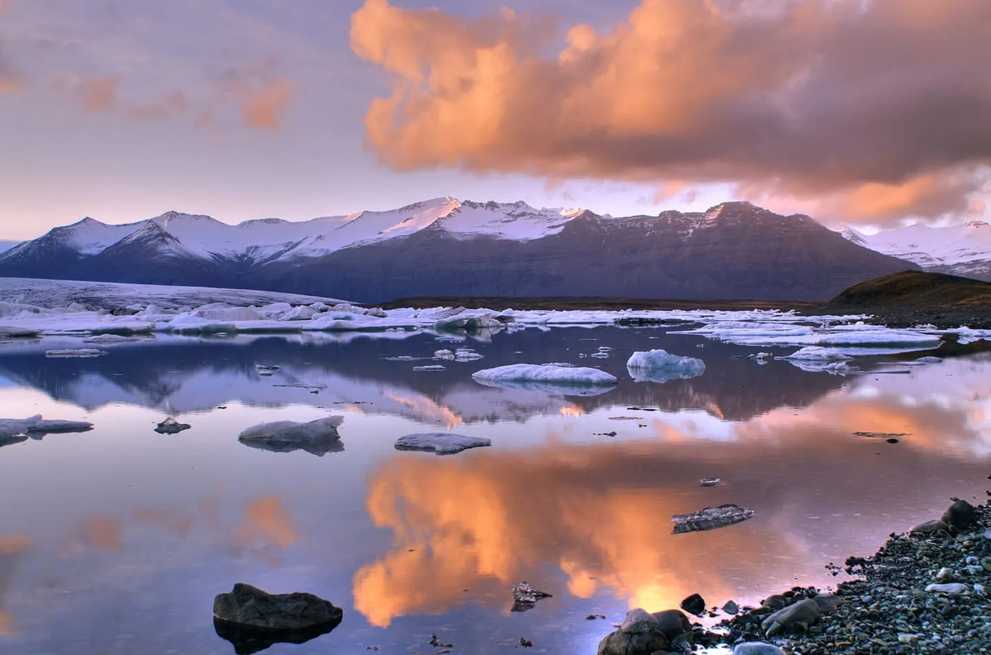 Icebergs floating in the still Jökulsárlón lagoon under a pink and orange sunset sky, with snowy mountains behind