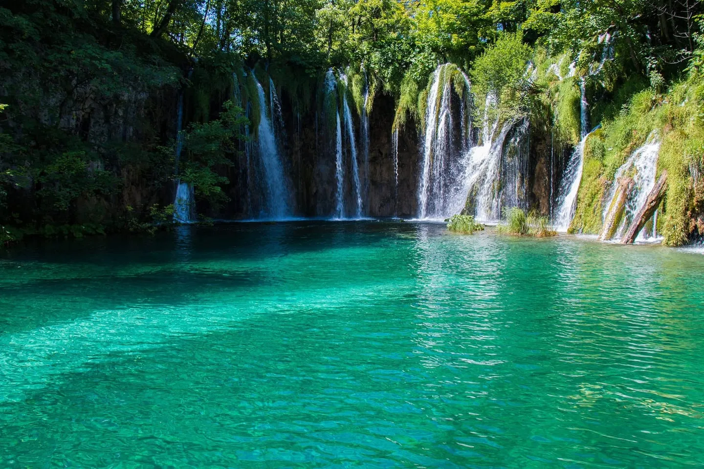 Turquoise water at Plitvice Lakes with a row of waterfalls pouring over mossy cliffs