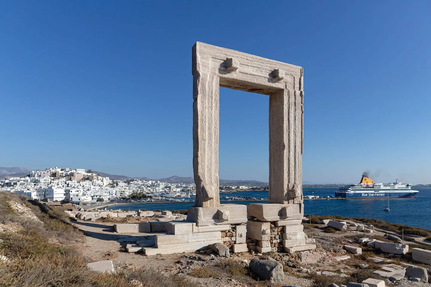 The Portara of Naxos, a huge marble doorway on a rocky islet, with the white town of Chora and a ferry in the harbor behind it